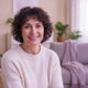 A candid portrait of a smiling middle-aged woman with dark, curly hair, wearing a cream sweater, in a softly lit living room 